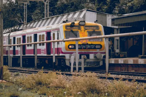 Front view of an Indian train at a railway station platform surrounded by greenery and infrastructure.