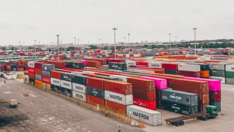 Press Note 3 (2026 Series): The - Colorful shipping containers stacked at a bustling port showcasing global trade.